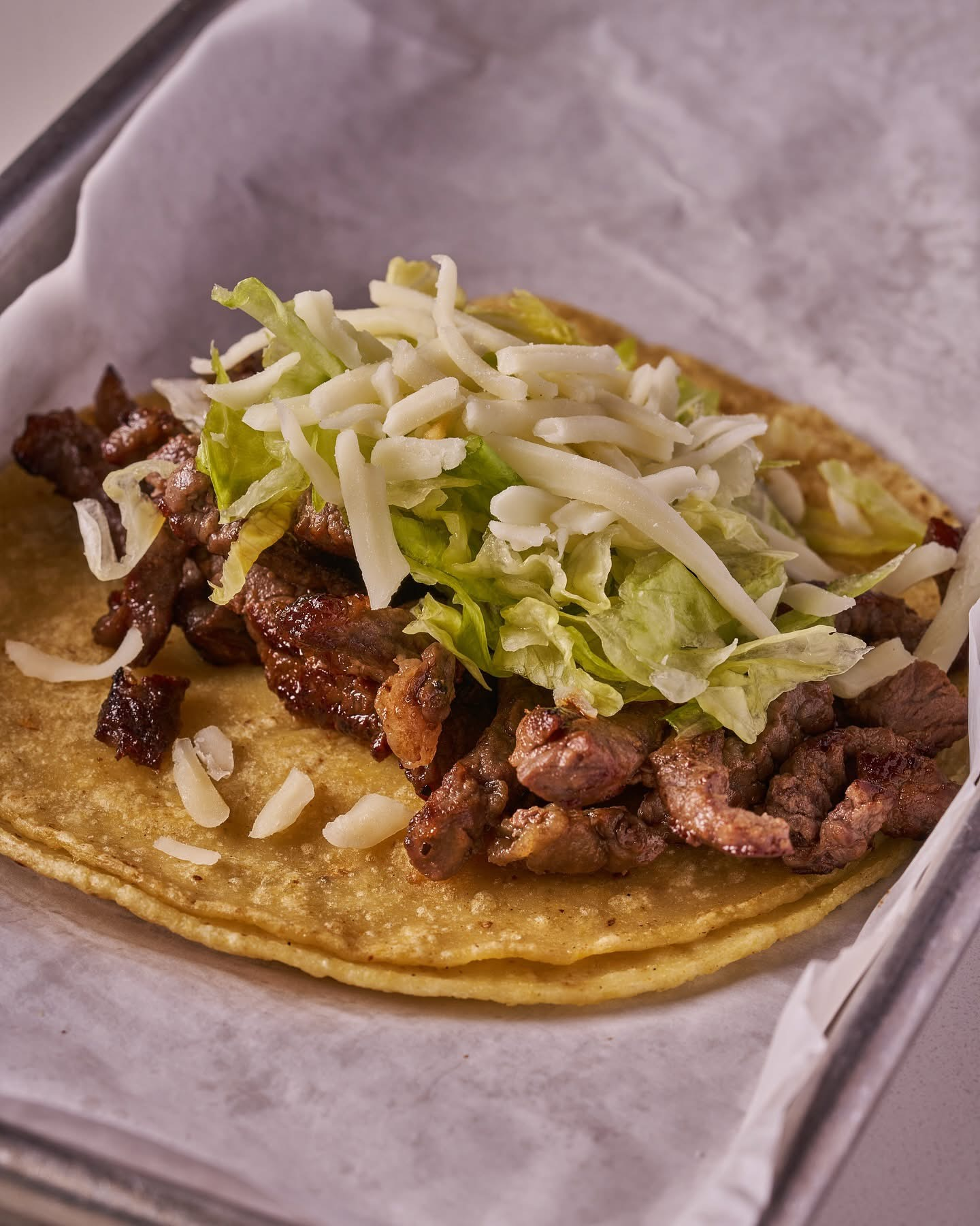 Taco with grilled meat, shredded lettuce, and cheese on a corn tortilla, in a paper-lined tray.
