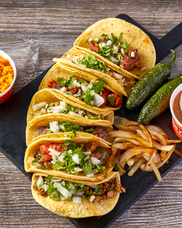 Tacos with salsa, onions, peppers, fries, and dipping sauces on a black serving board.