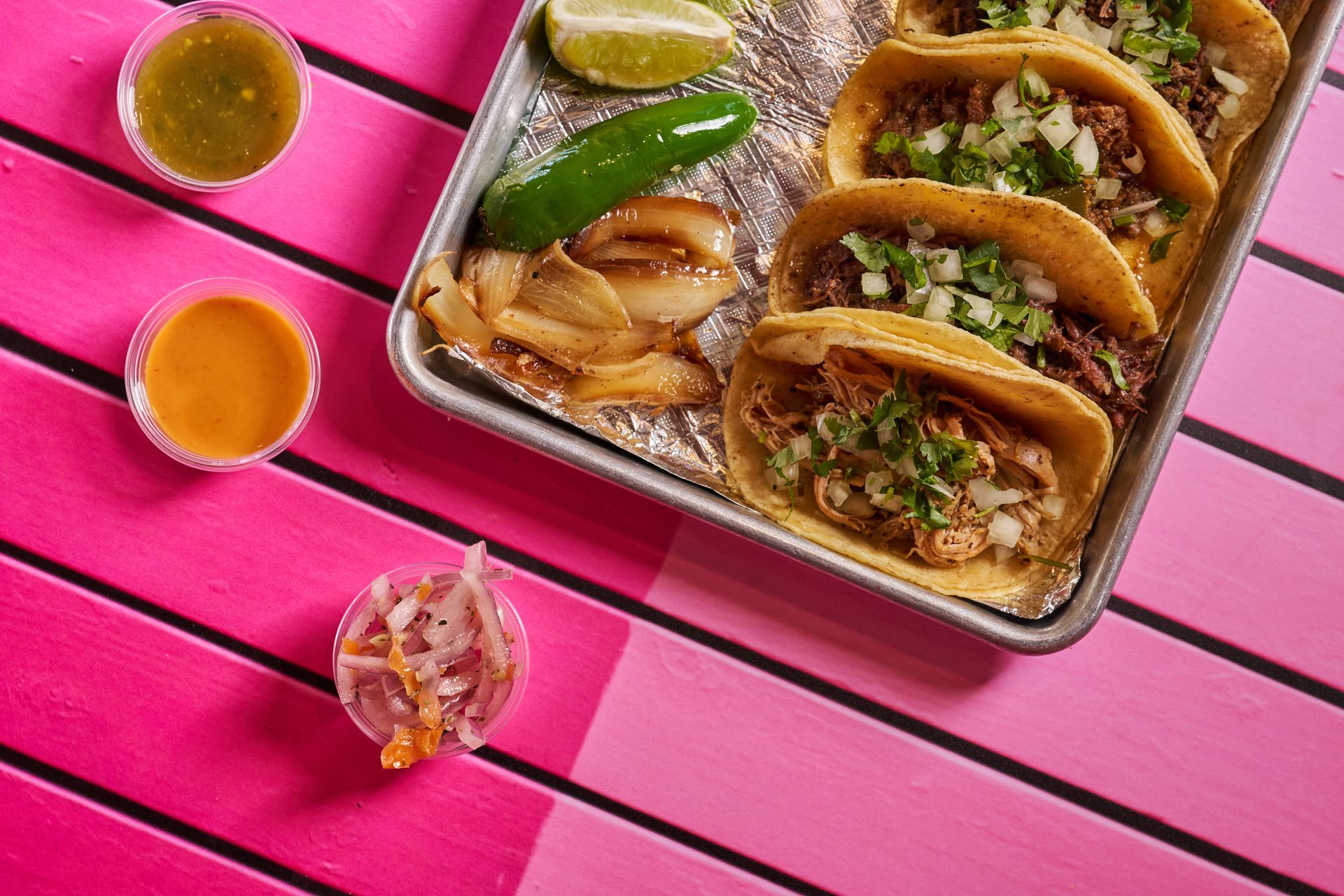 Tacos on a tray with toppings and sauces on a pink and white striped surface.