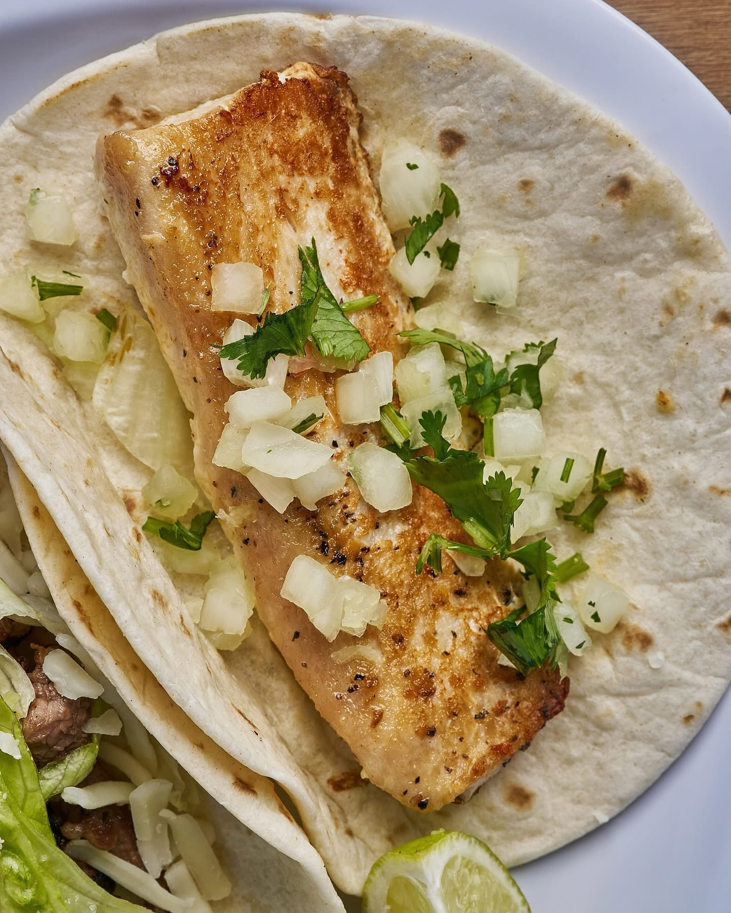 Fish taco with grilled fish, onion salsa, cilantro, and lime on a white plate.