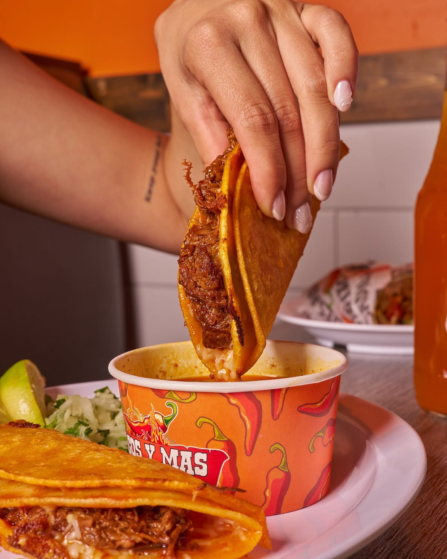 Hand dipping a taco into a consommé. Plate with taco, lime, and slaw. Orange cup has red chili peppers.