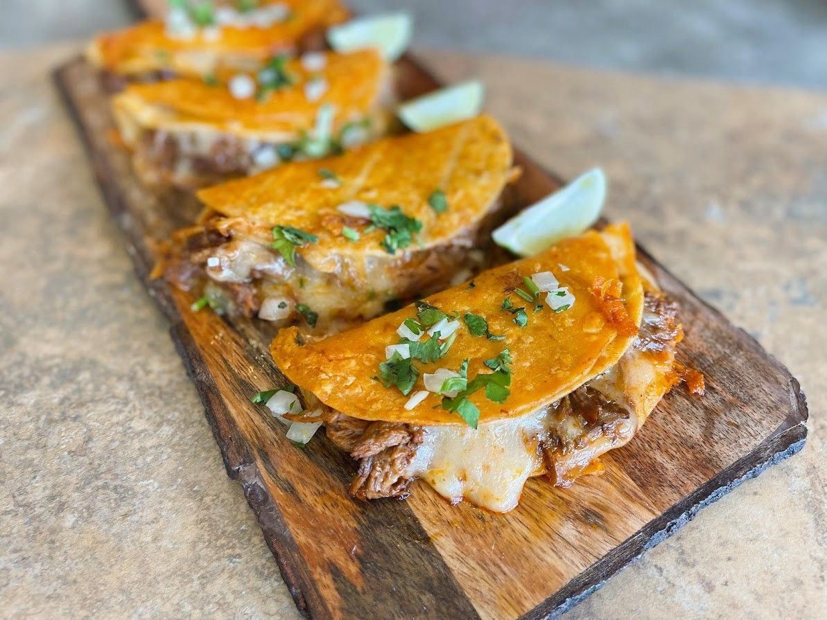 Birria tacos on a wooden board, garnished with cilantro, onions, and lime wedges.