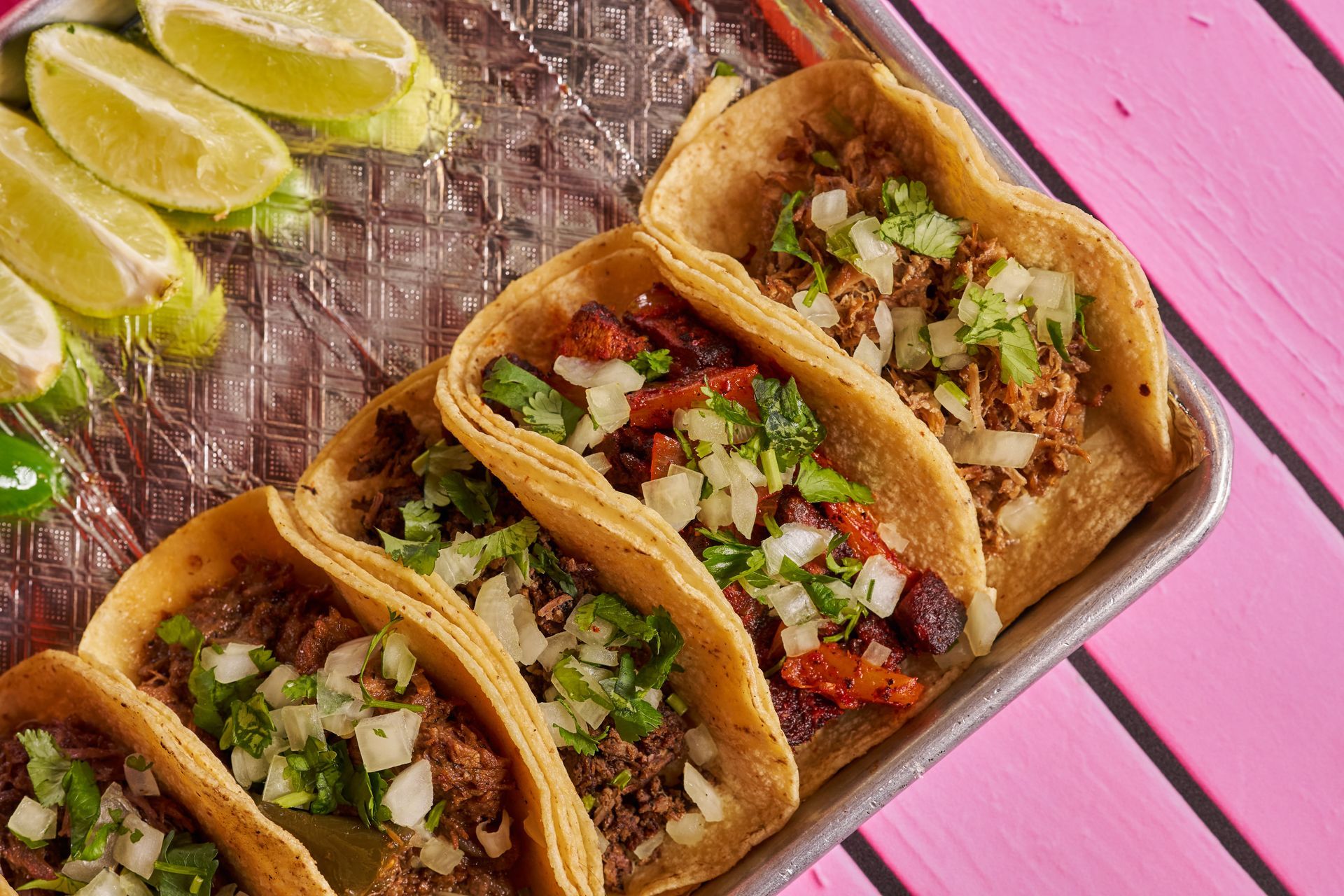 Tacos in hard shells, topped with onions and cilantro, with lime wedges on a tray.