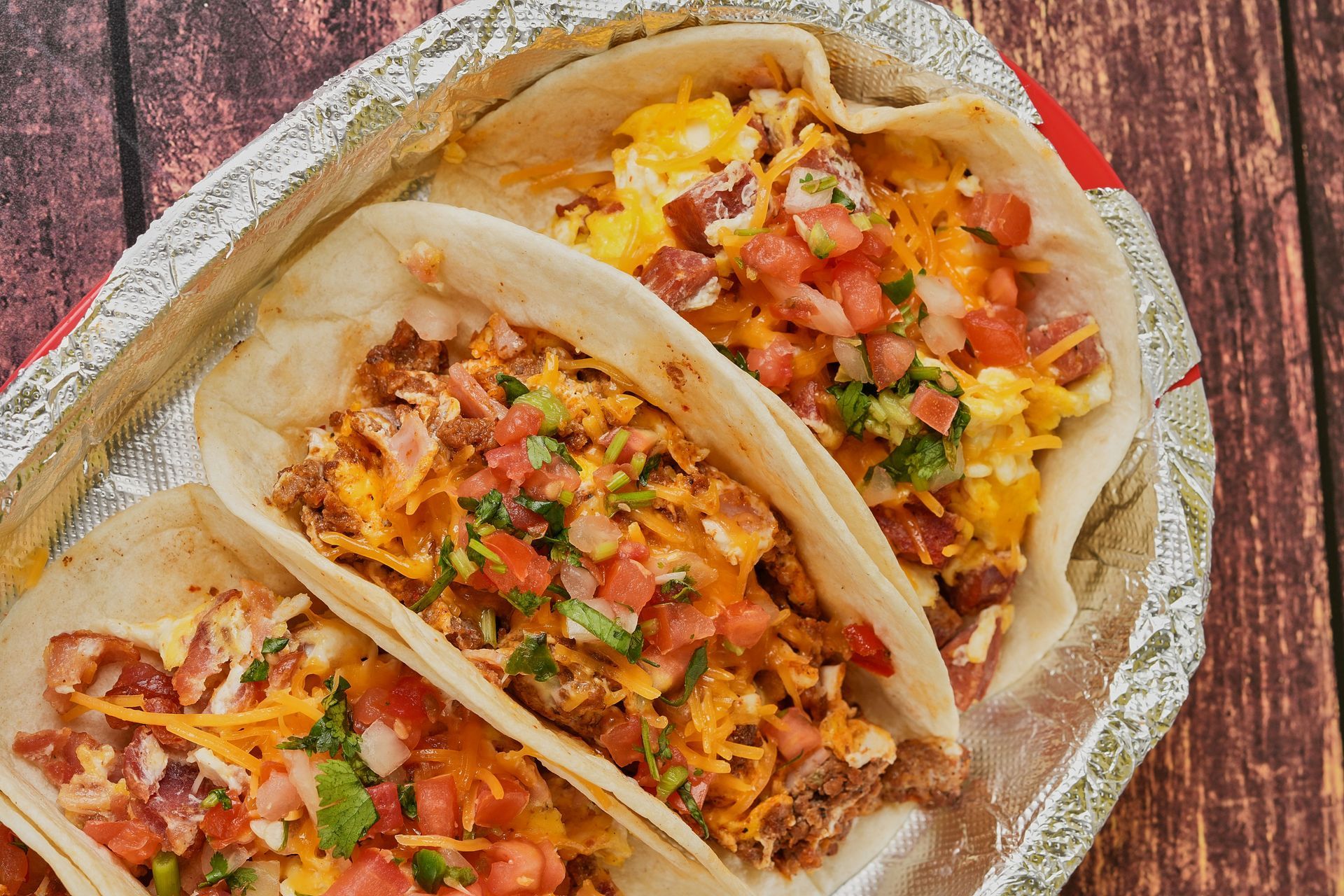 Breakfast tacos with scrambled eggs, meat, cheese, and pico de gallo, in a foil-lined tray.