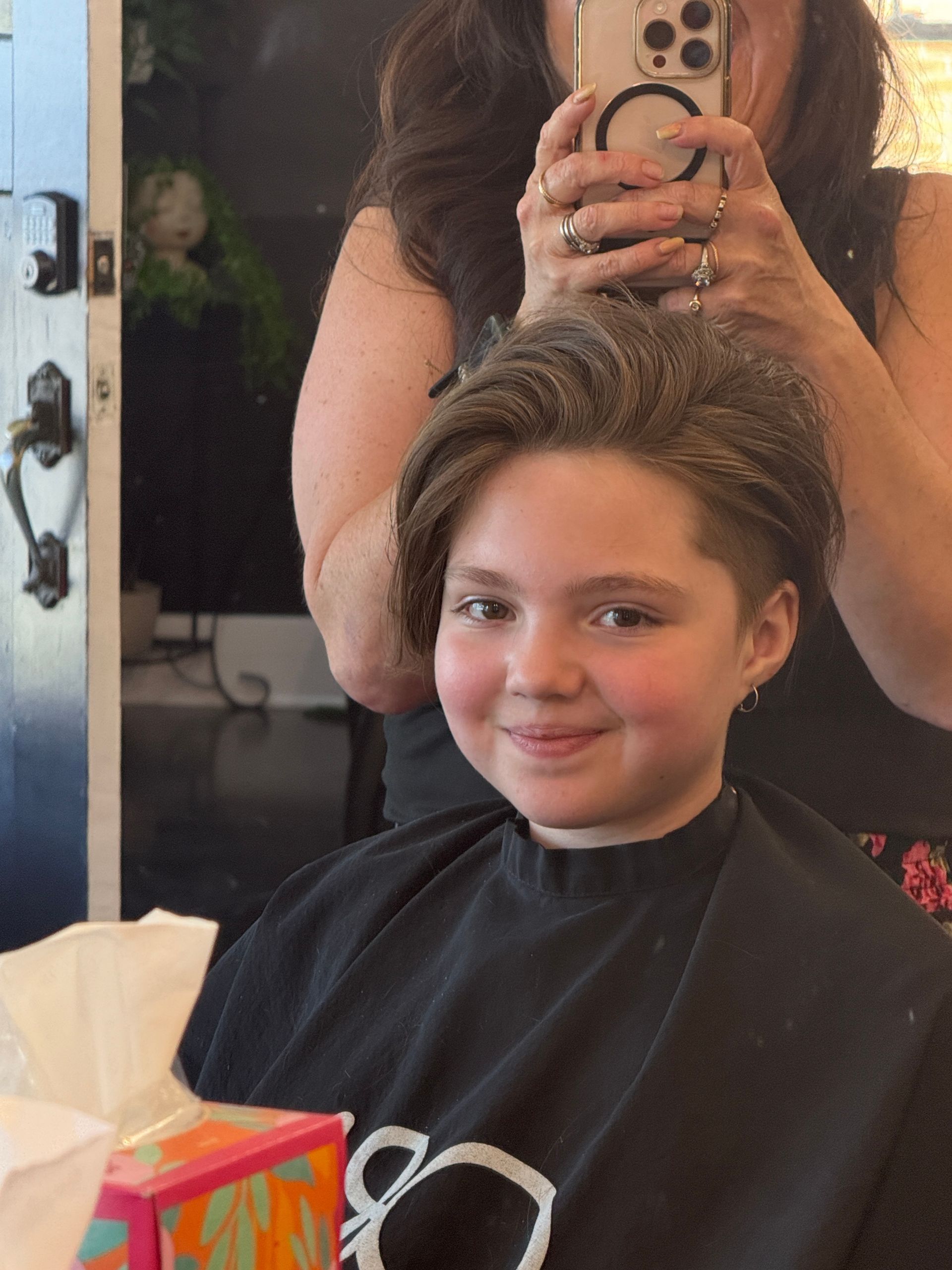 Person smiling in a salon chair while getting a haircut, with a stylist visible in the mirror.