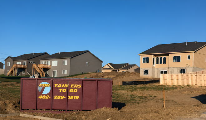 Residential houses behind a maroon dumpster in a grassy construction lot under a clear blue sky.