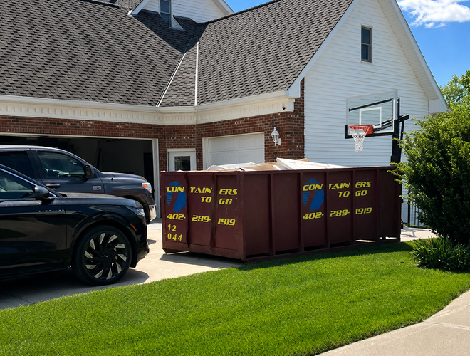 Large brown dumpster in a suburban driveway beside a black SUV and a basketball hoop