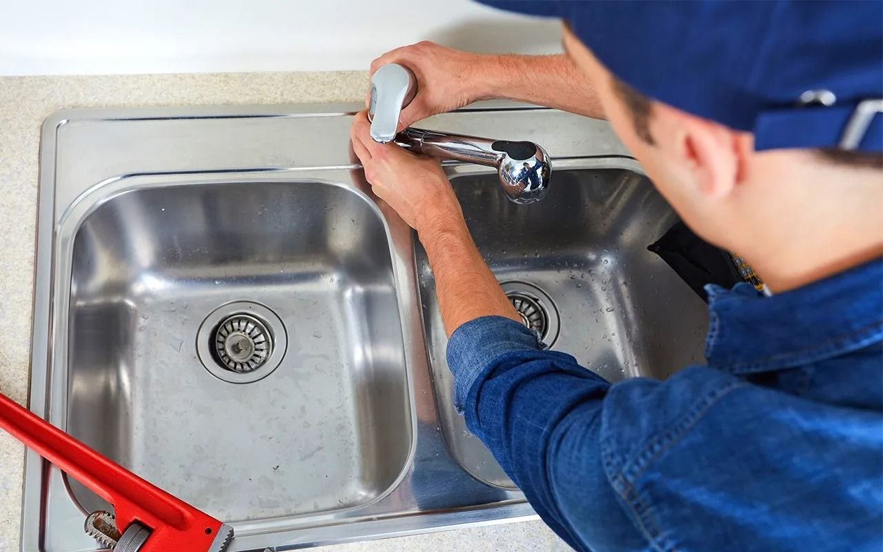 Plumber in blue uniform working on a kitchen sink with a red wrench, indoors.
