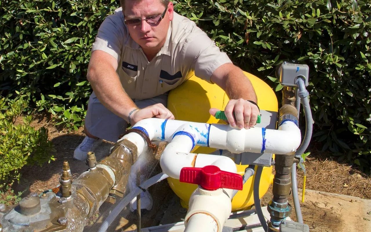 Man in uniform inspecting water pipes; white and red pipes, yellow tank, water leaking. Outdoor setting.