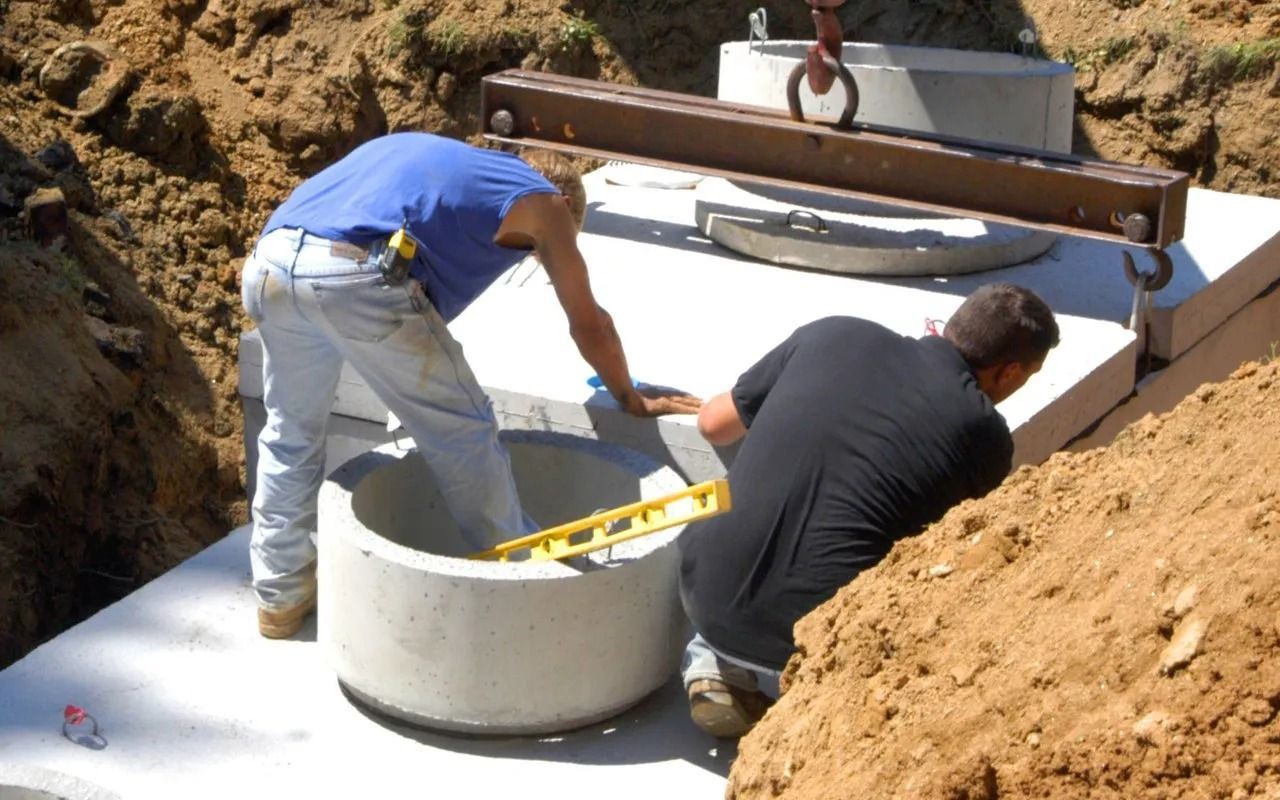 Two workers installing a concrete well ring in a dirt excavation. One uses a level.