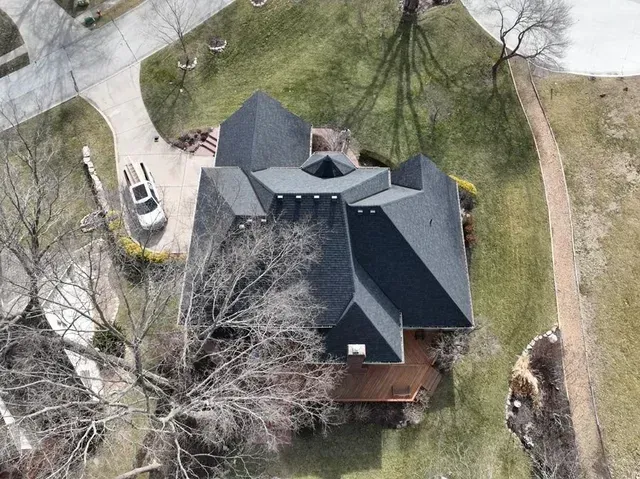 Aerial view of a dark-roofed house with a car in the driveway and bare trees, on a grassy area.