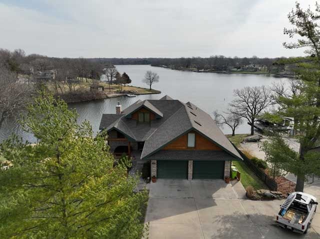 House with brown roof and green garage doors on lake, trees surrounding.