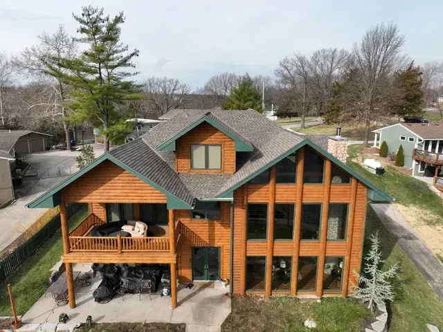 Two-story wooden house with large windows, green trim, and a porch, on a grassy lot.