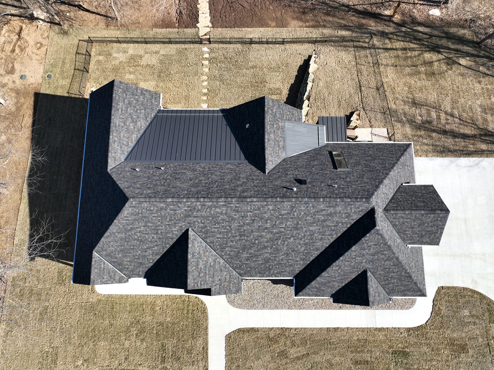 Overhead view of a house with a dark gray shingled roof, casting shadows on the brown grass.