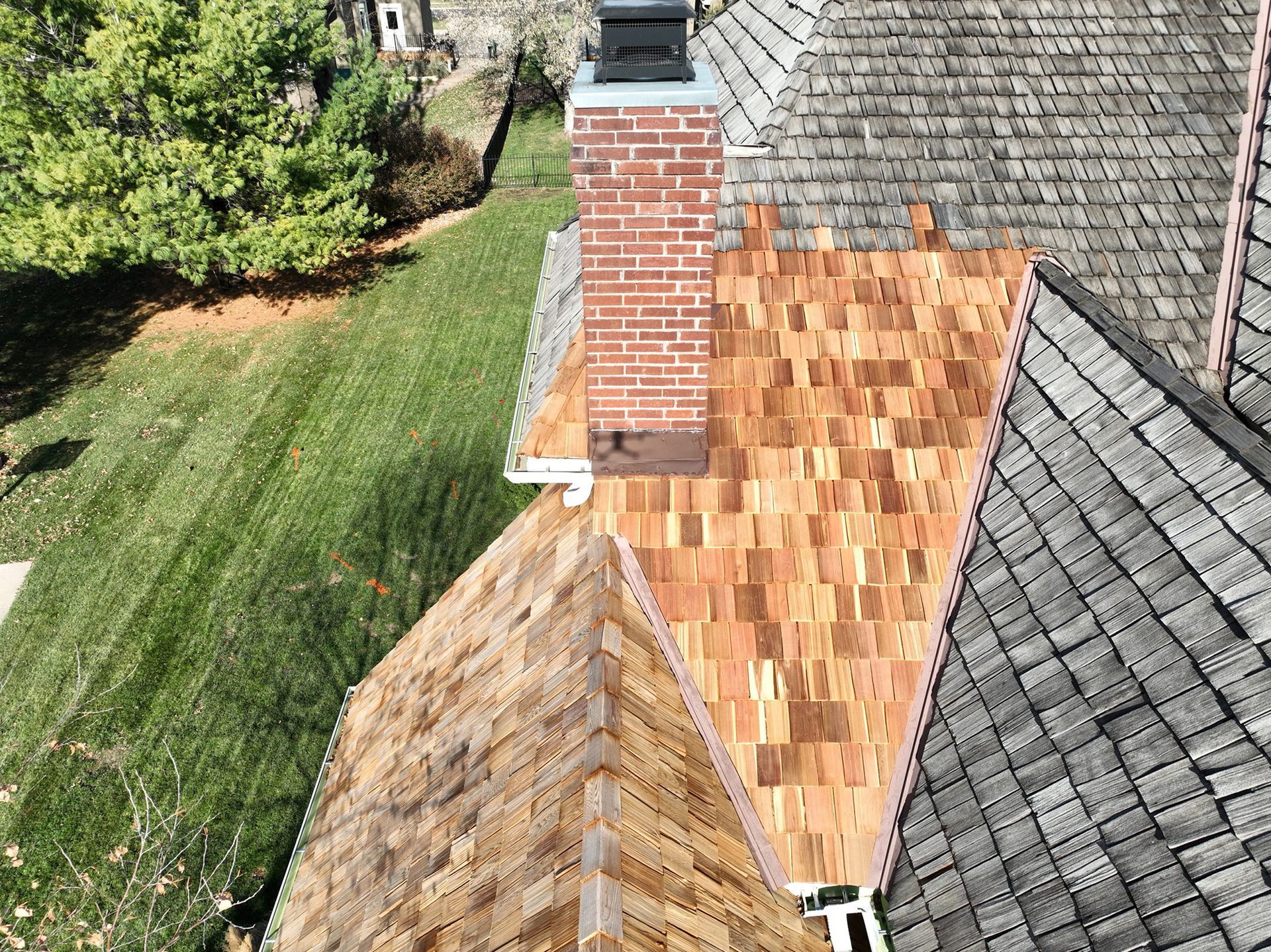 Overhead view of a roof with a brick chimney and new wood shingles next to older gray shingles.
