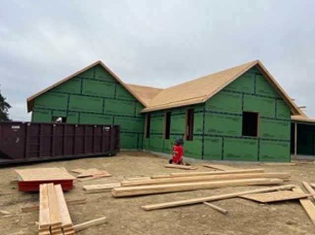 House under construction, green sheathing, wood framing, brown roof, lumber and a dumpster on site.