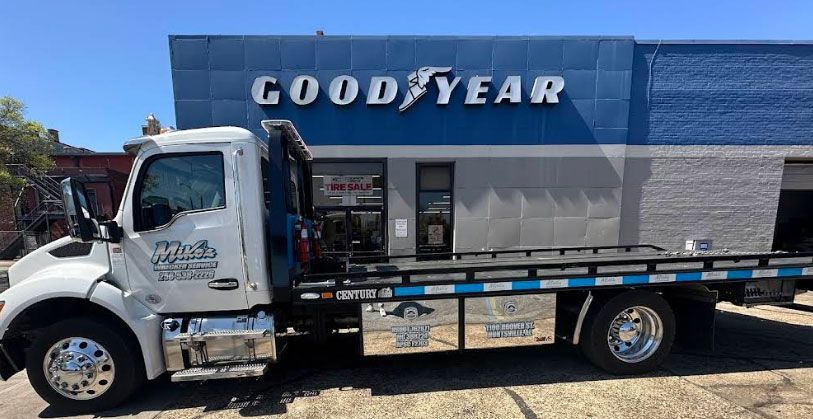 White tow truck in front of a Goodyear tire store, blue and white building, sunny day.