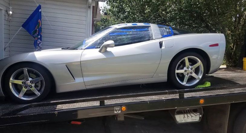 Silver Corvette sports car on a tow truck, parked near a house with a blue flag.