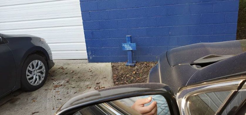 A car's side mirror reflects a hand, near a blue wall and another parked car.