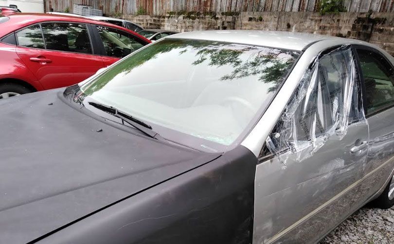 Silver car with plastic covering the side and windshield; a red car is partially visible in the background.