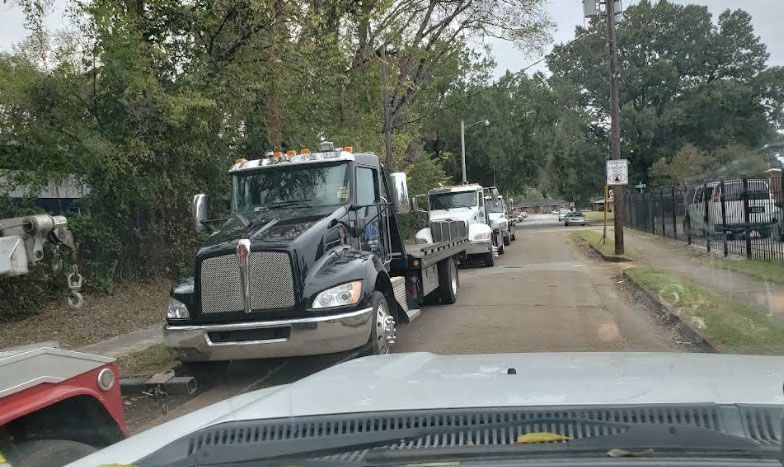 Black tow truck with a white truck on its flatbed parked on a street near trees and a building.