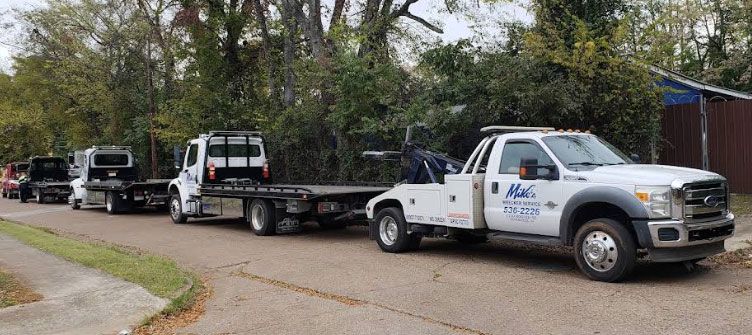 A line of tow trucks parked on a road beside green trees and a building.