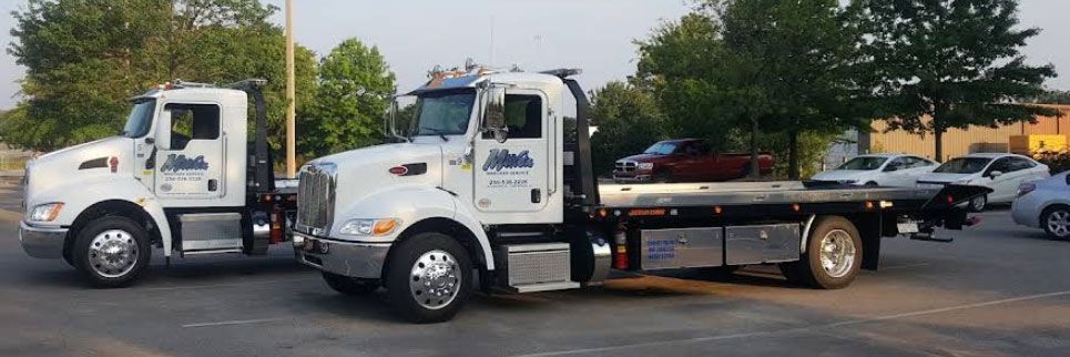 Two white tow trucks, one loaded with a maroon car, in a parking lot. Trees and other cars are visible.