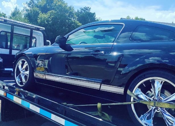 Black Shelby Mustang on a tow truck. Chrome wheels, white stripe, bright sunlight, and blue sky.