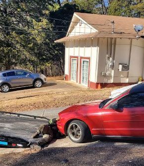Red car crashed; person on the ground next to it. Grey SUV and a small white building are in the background.