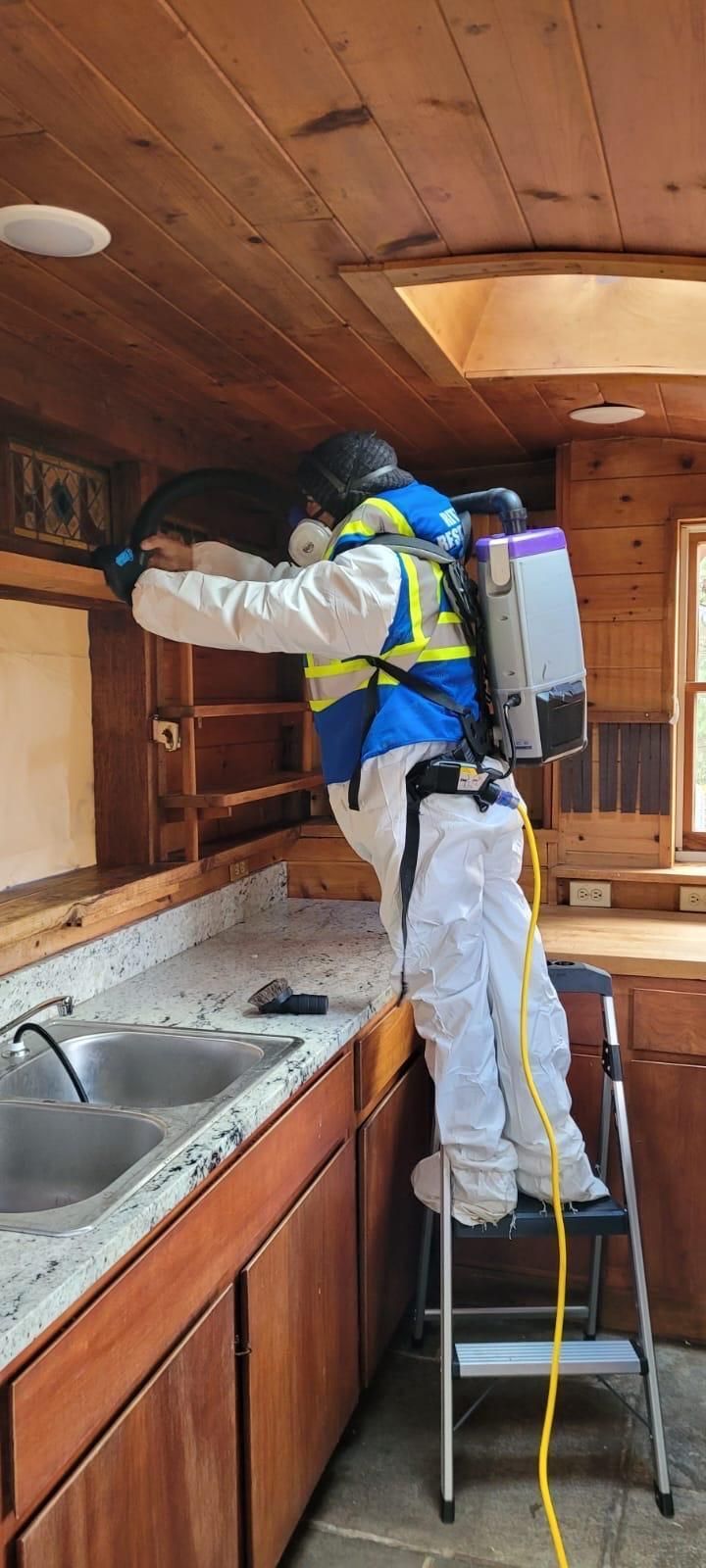 Worker in white coveralls cleans a wooden kitchen ceiling while standing on a ladder with a backpack sprayer.