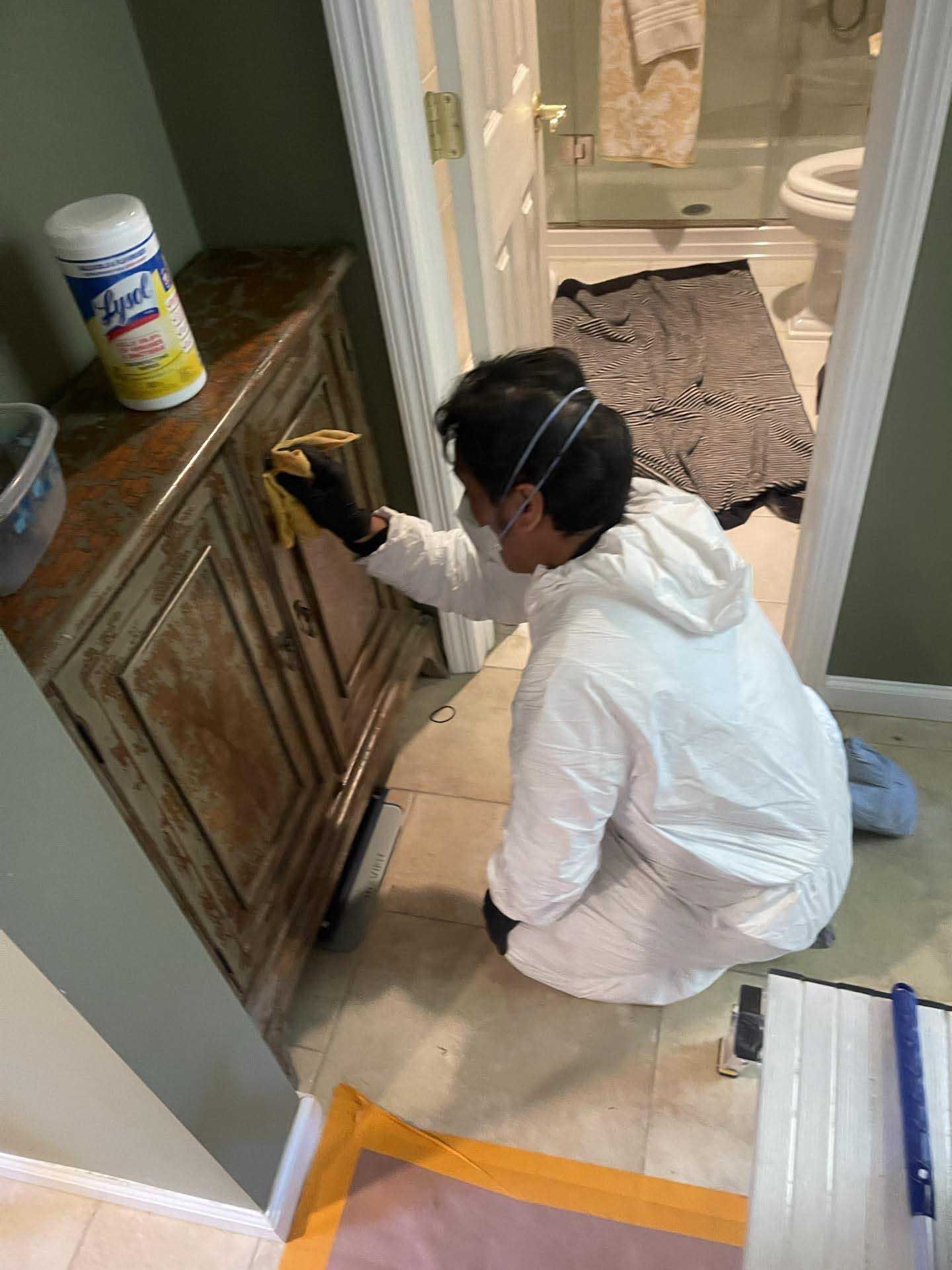 A person in a white hazmat suit and mask cleans a wooden cabinet in a bathroom with a disinfectant wipe.