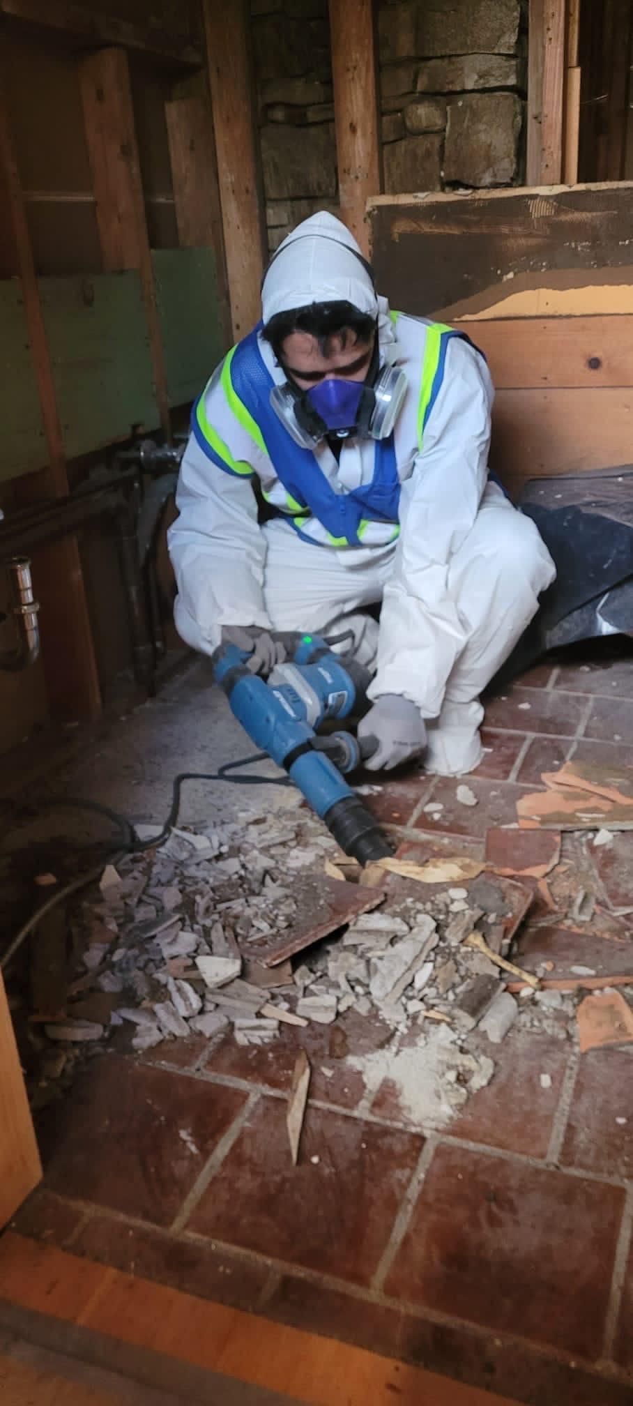 Worker in white protective gear using a jackhammer to break a tiled floor indoors.