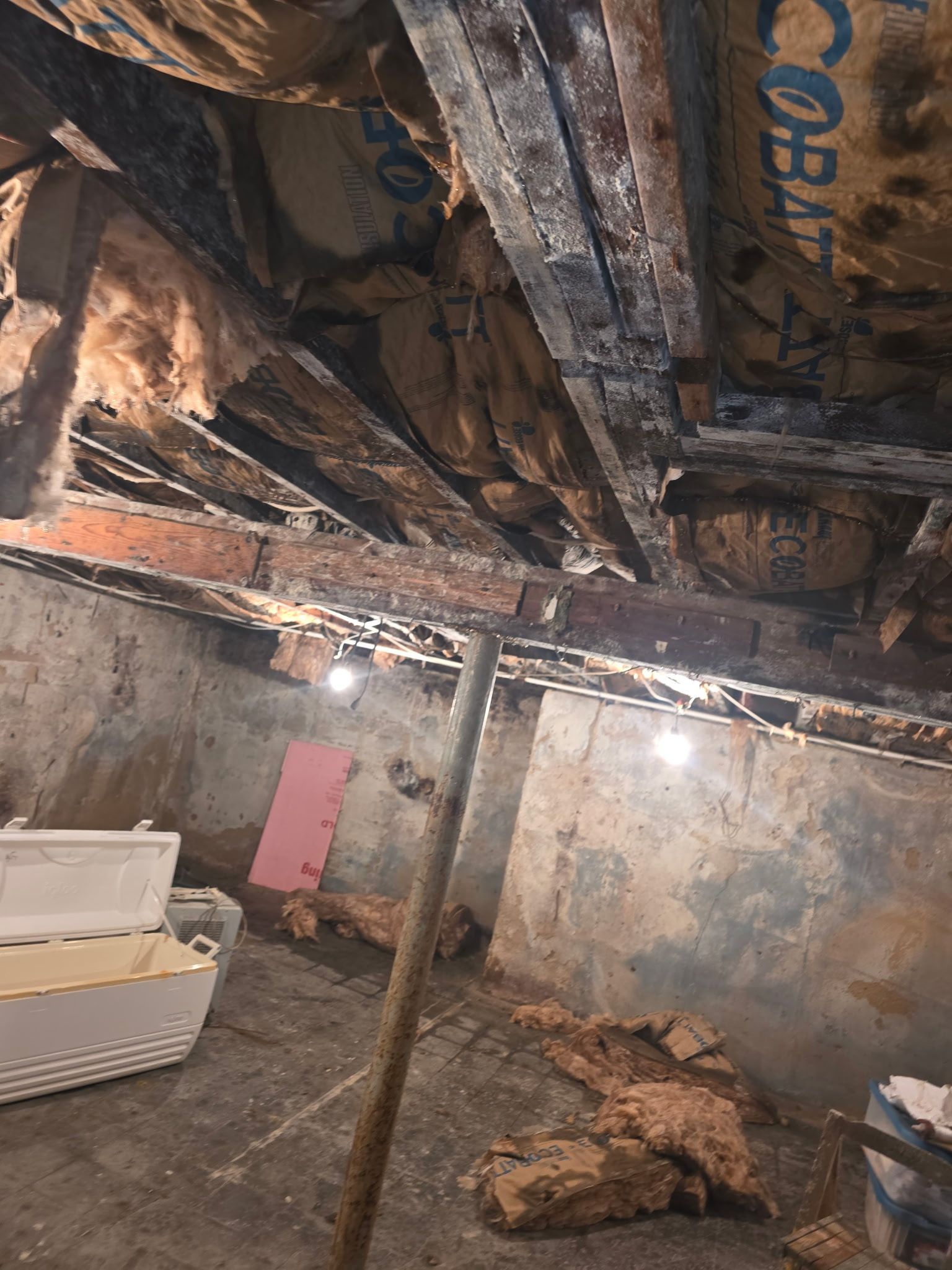 Damaged basement ceiling with exposed beams, cracked walls, and debris on the floor, with a white couch below.