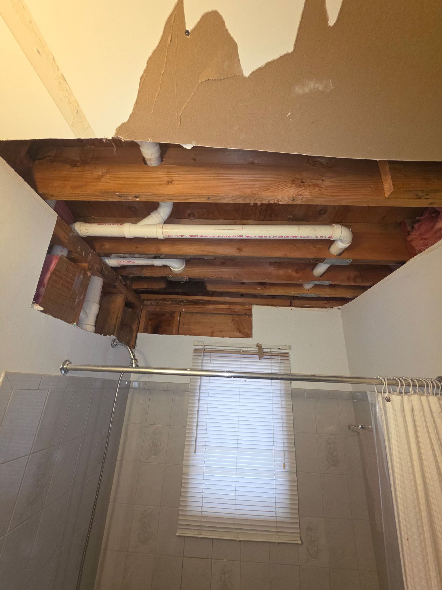 Bathroom ceiling with exposed wooden beams, pipes, and a small window above a shower curtain.