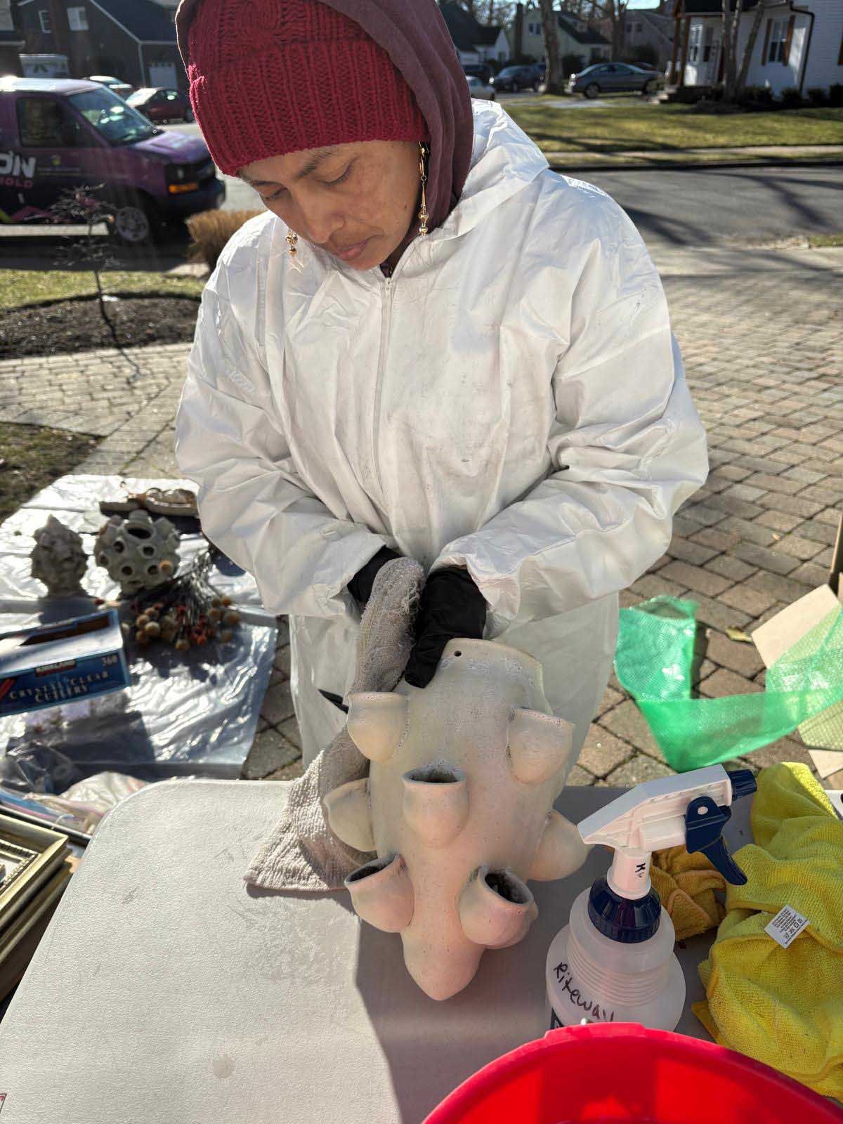 A person in a white hazmat suit and red hat cleans a ceramic strawberry planter on an outdoor table.
