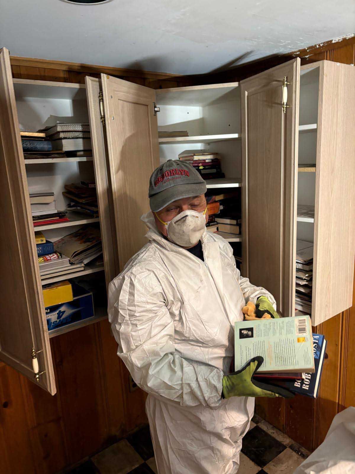 A person in a white hazmat suit, mask, and cap sorts books from shelves in a room with wood-paneled walls.