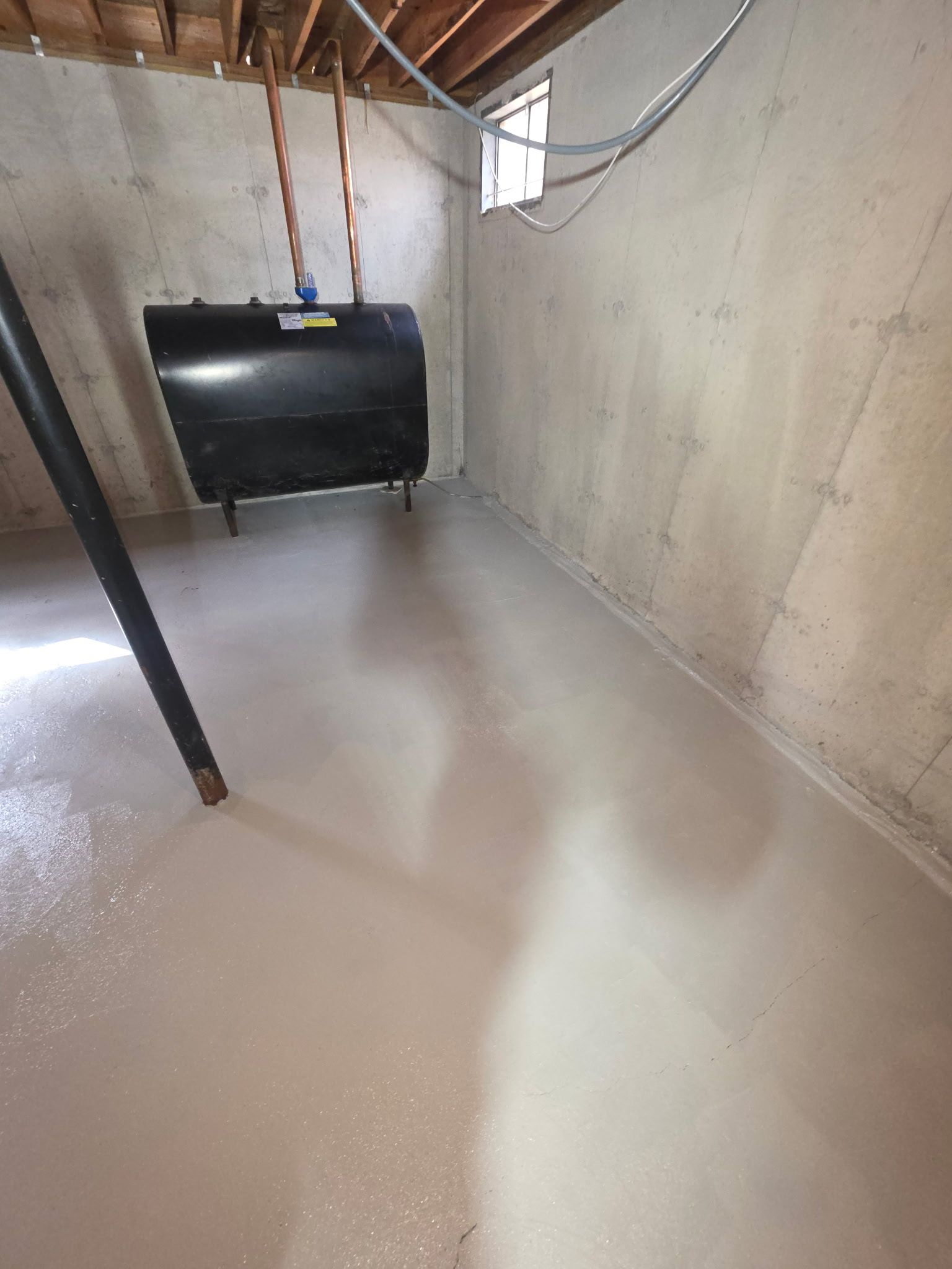 Basement with freshly coated glossy floor, unfinished concrete walls, exposed pipes, and a black tank on the far wall