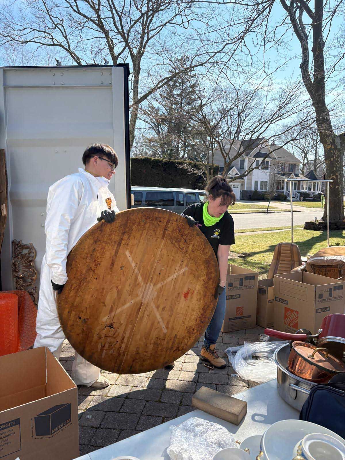 Two people in casual and protective clothing lift a large, round wooden tabletop near an open moving container outdoors.