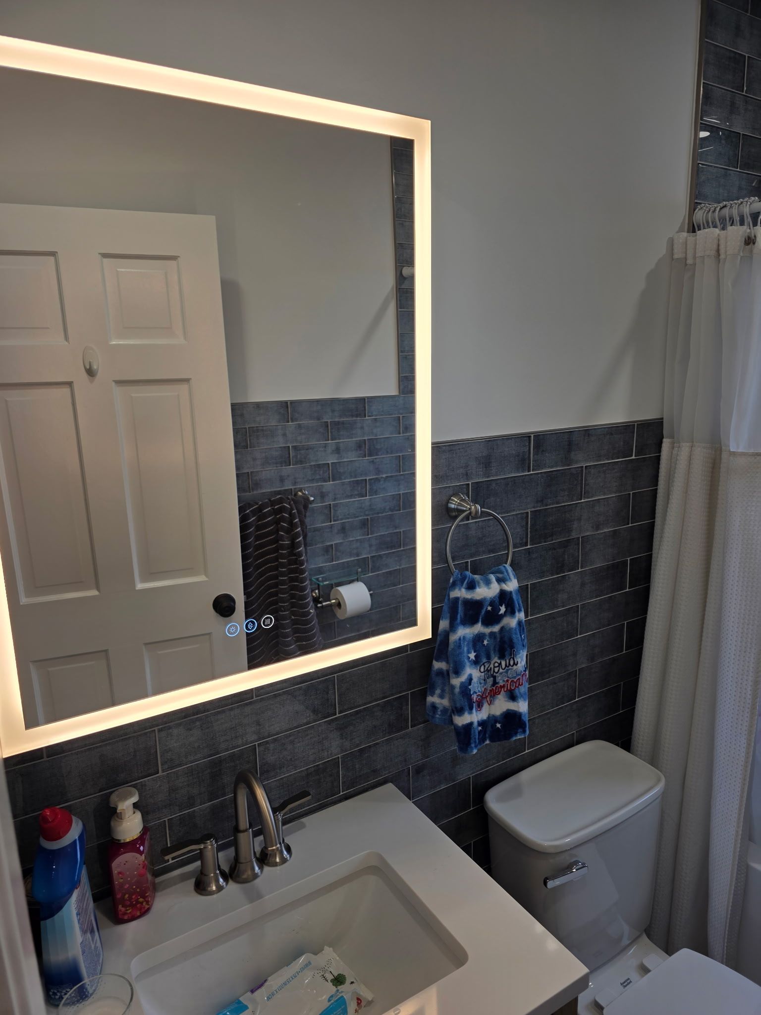 Small bathroom with lit mirror above sink, tiled shower wall, toilet, and window curtain.