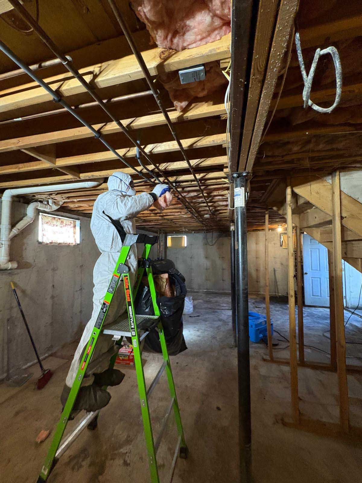 A person in protective coveralls stands on a ladder, working on electrical wiring in an unfinished basement.