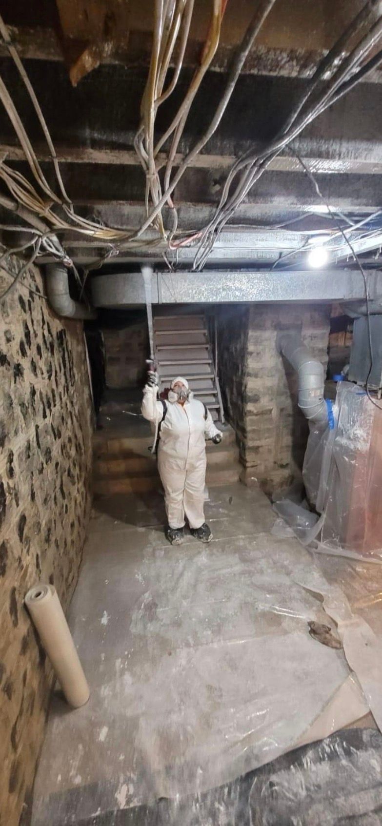 Person in white coveralls standing in a dim basement with exposed pipes and brick walls