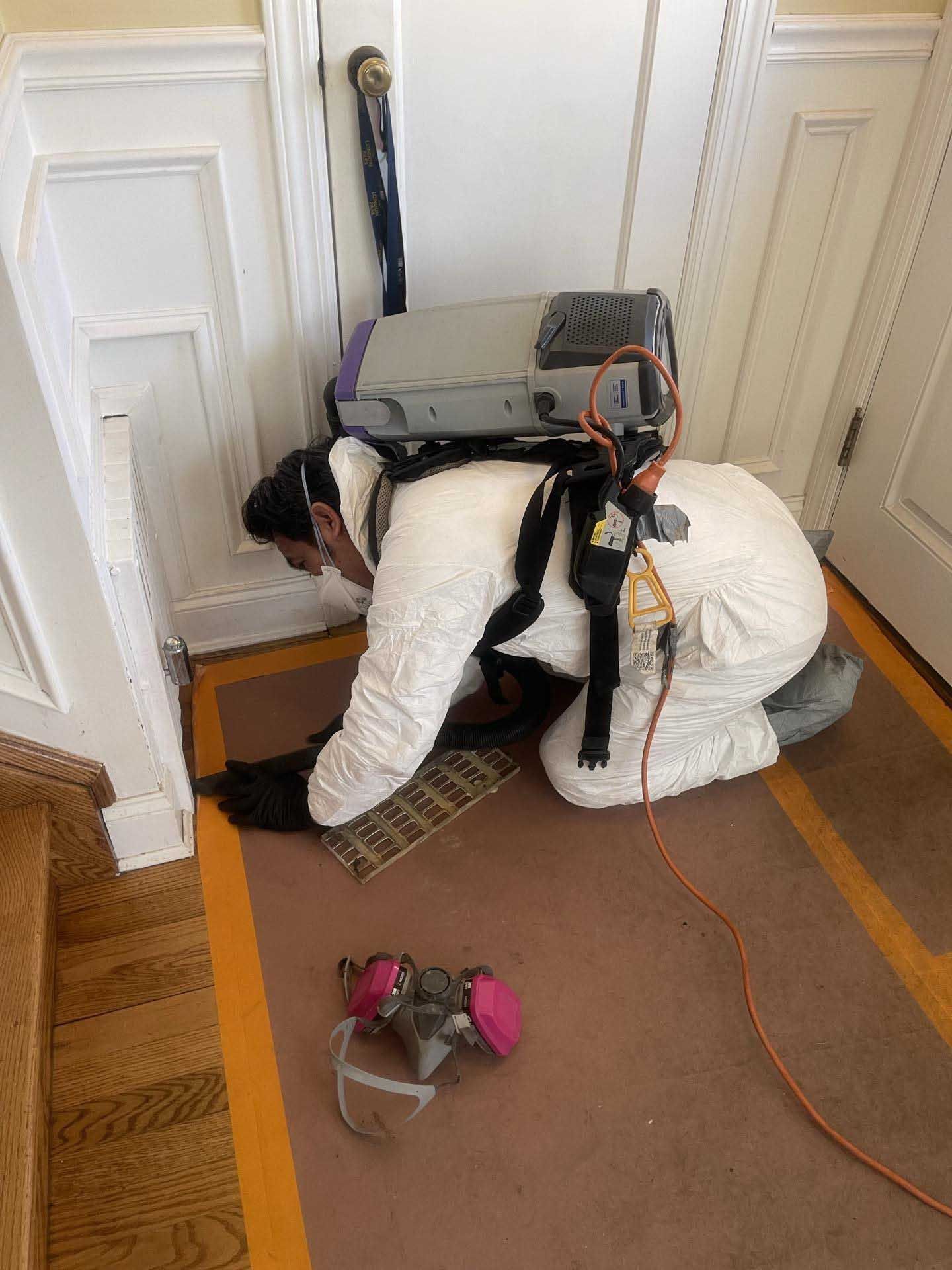 A person in a white protective suit and backpack vacuum works on the floor near a vent, with a respirator on the rug.
