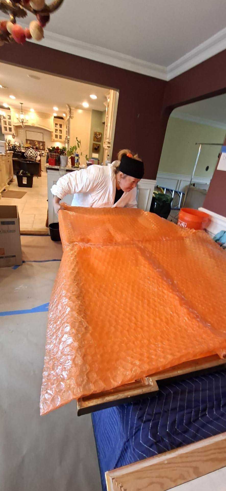 A person in a white jacket leans over a large wooden surface covered with orange bubble wrap in a home interior.