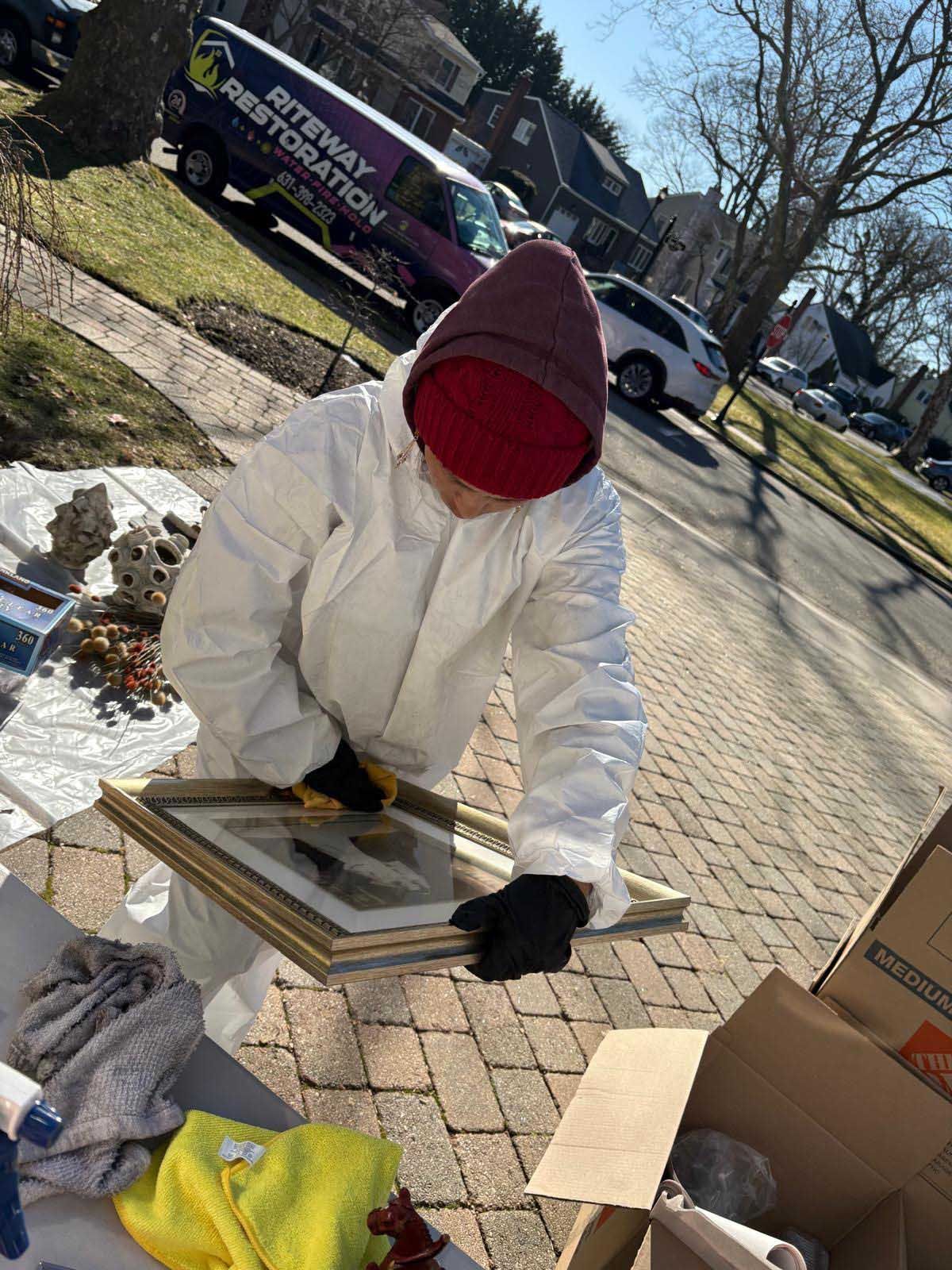 A person in white protective gear and a red beanie cleans a framed picture outdoors near a Riteway Restoration van.