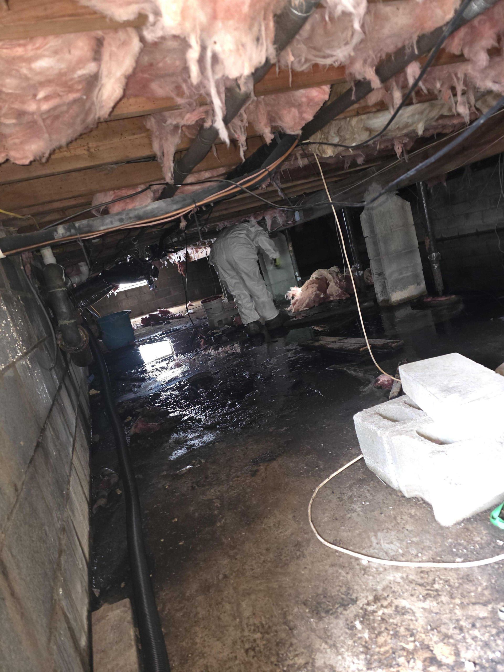 A person in protective gear works in a dark, damp crawl space with exposed insulation and standing water on the ground.