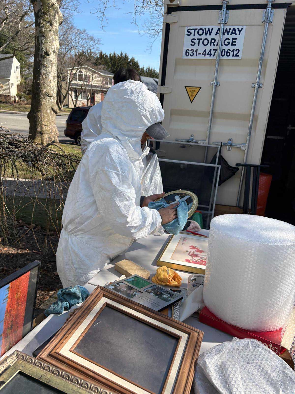 A person in a white hazmat suit cleans items at a table outdoors in front of a Stow Away Storage container.