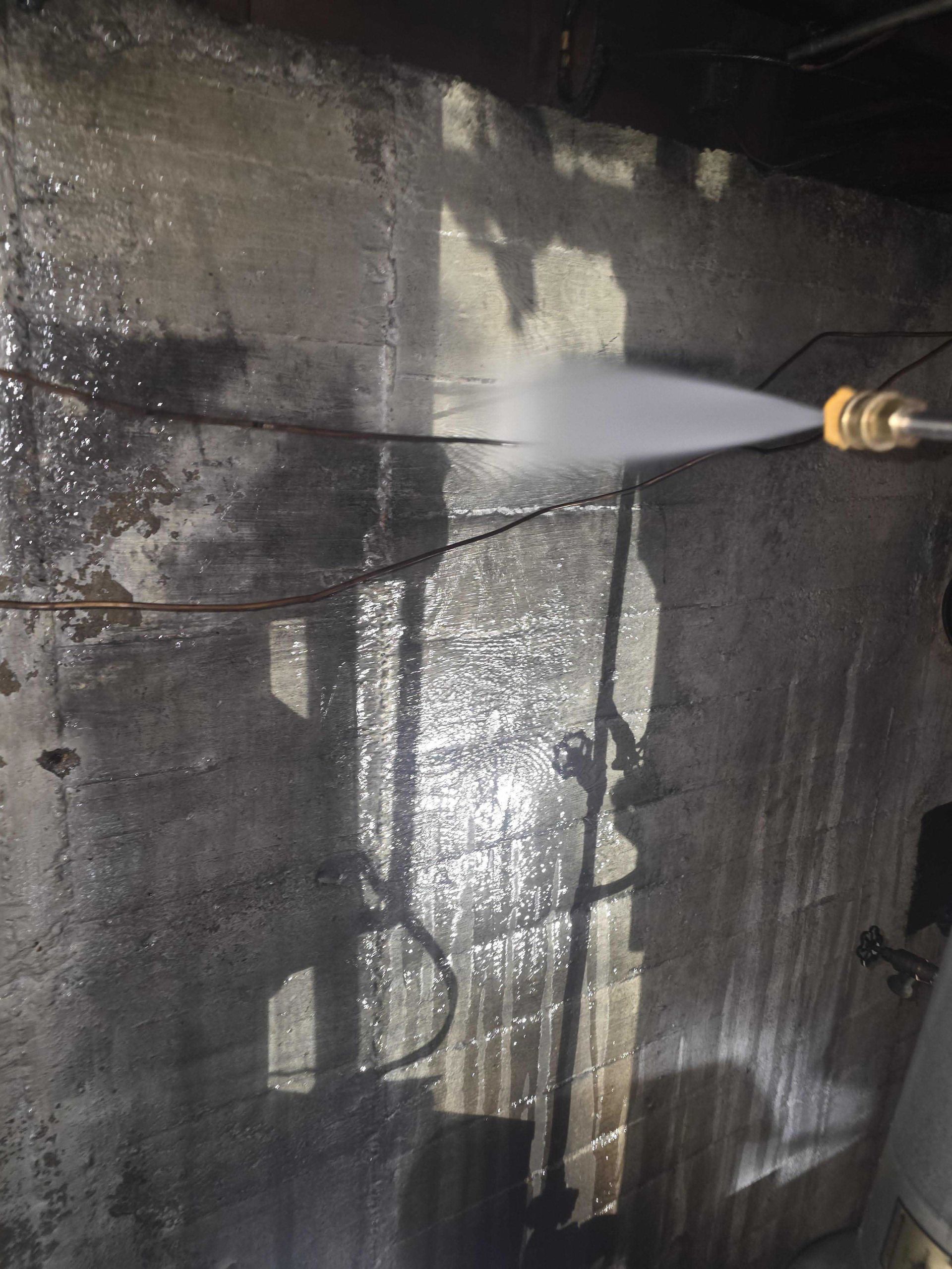 A power washer sprays water onto a dark, stained concrete wall to clean it.