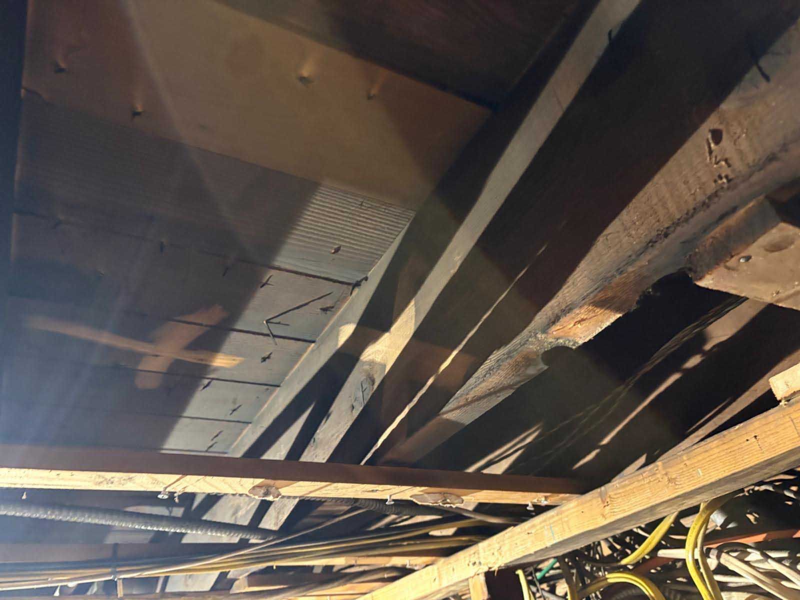 An upward view of exposed ceiling joists, wooden framing, and electrical wiring in an unfinished attic space.