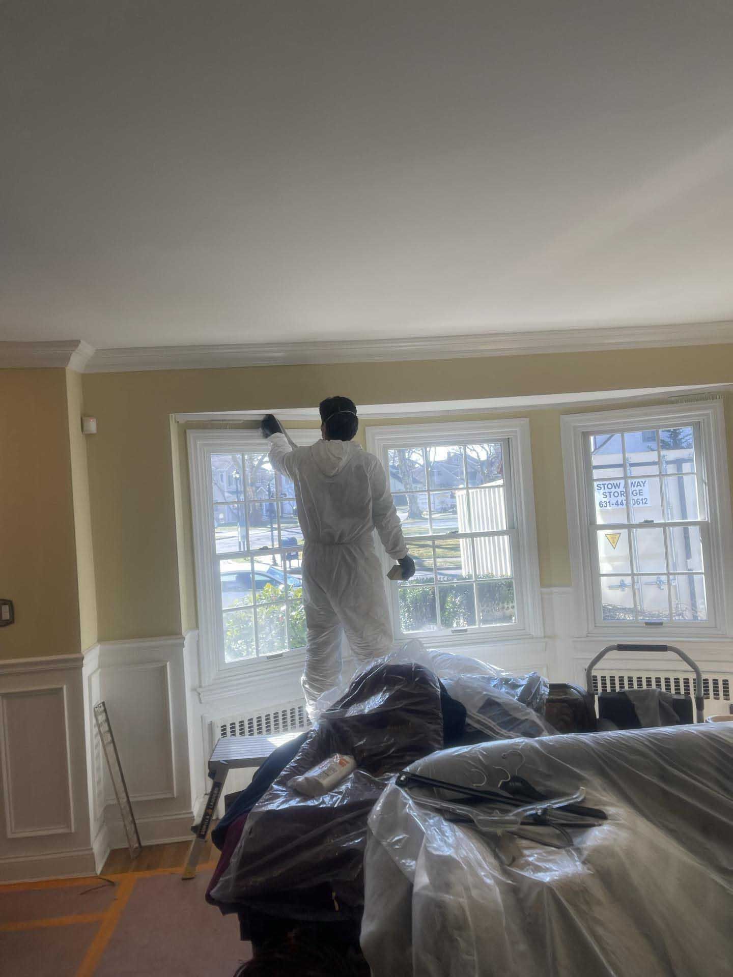 A person in protective coveralls paints the trim above windows in a room with light yellow walls.