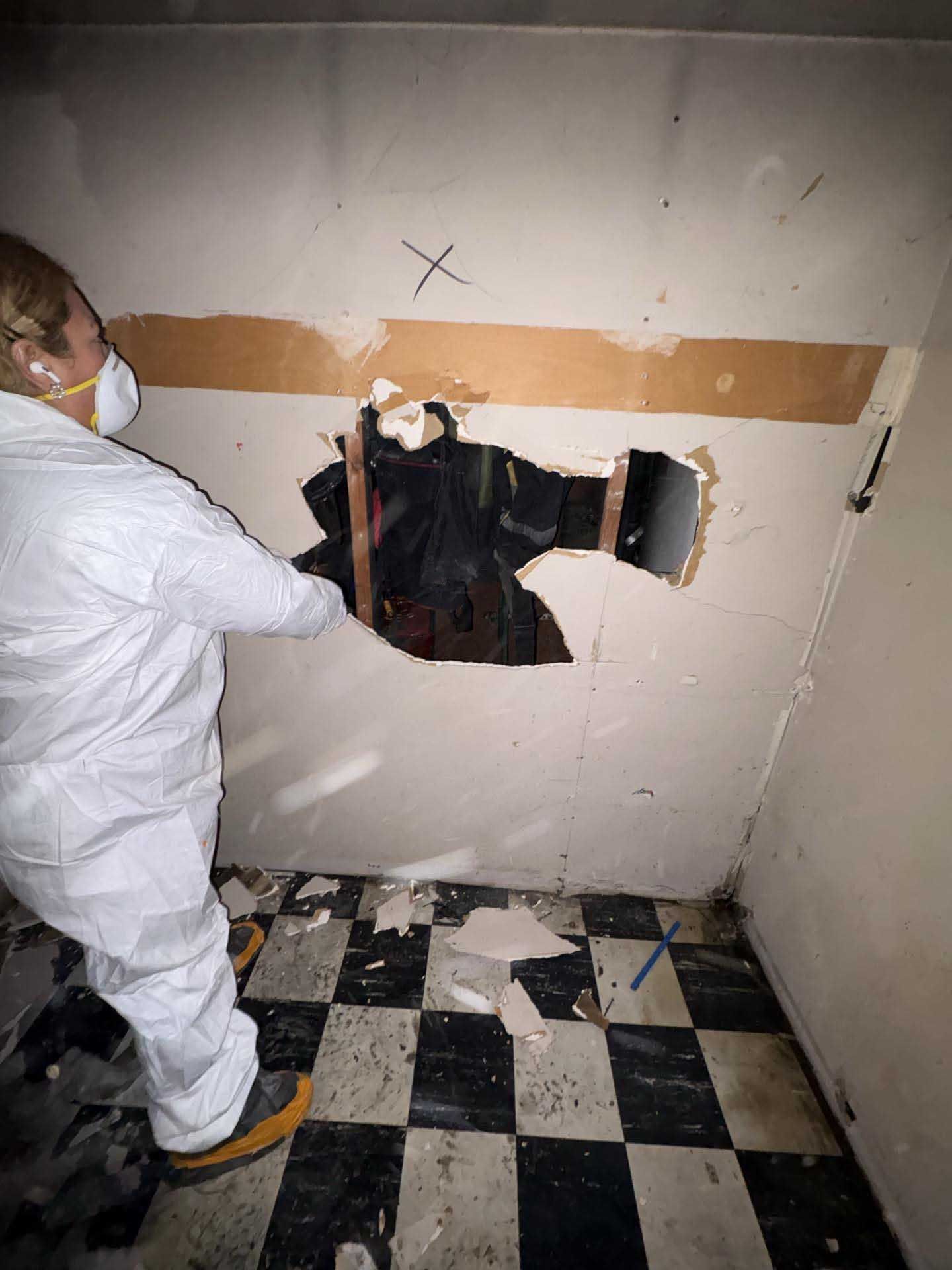 A person in a white hazmat suit and mask inspects a hole in a wall above a black-and-white checkered floor.